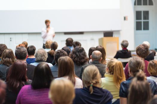 Female speaker giving presentation in lecture hall at university workshop. Audience in the conference hall. Rear view of unrecognized participant in audience. Scientific conference event.