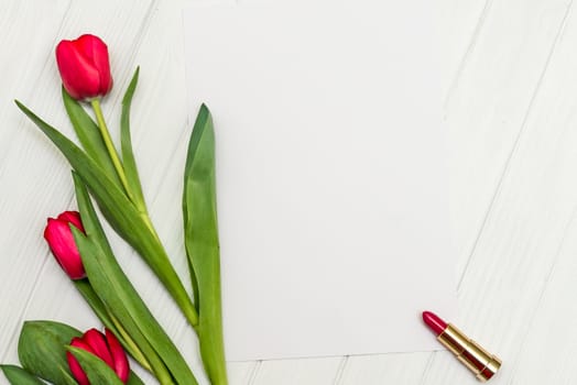 red tulips, lipstick and sheet of paper for your greetings on the background of white wooden board