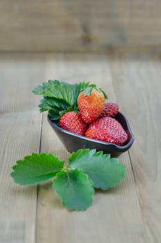 fresh strawberry fruits in black bowl with green leaves over wooden background