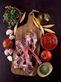 Raw Lamb Ribs with Tomatoes, Spices, Herbs, Olive Oil and Garlic on Wooden Cutting Board closeup on Dark Wooden background. Concept Ready to Roast