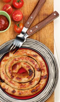Delicious Grilled Spiral Sausage on Striped Plate with Ketchup, Cherry Tomatoes  and Rustic Fork and Knife on Wooden Cutting Board closeup on Plank background