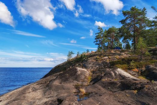 Blue tourist tent on a high rocky shore of Lake Ladoga on the background of blue sky with white clouds in Karelia.