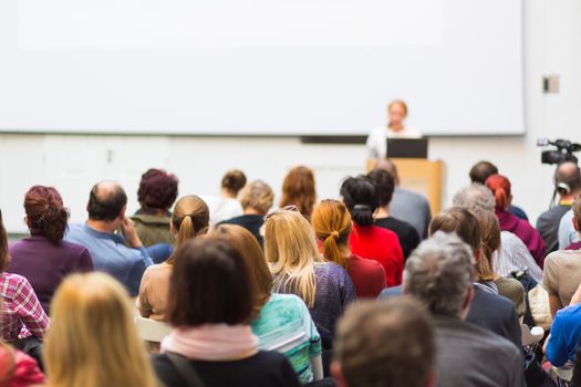 Business and entrepreneurship symposium. Female speaker giving a talk at business meeting. Audience in conference hall. Rear view of unrecognized participant in audience. Copy space on whitescreen.