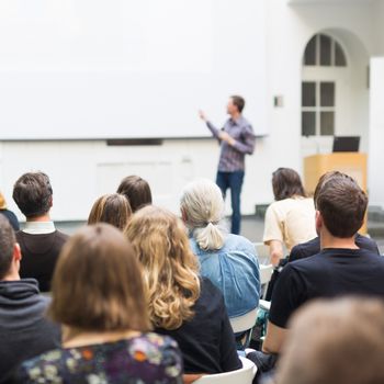 Male speaker giving presentation in lecture hall at university workshop. Audience in conference hall. Rear view of unrecognized participant. Scientific conference event. Copy space on whitescreen.