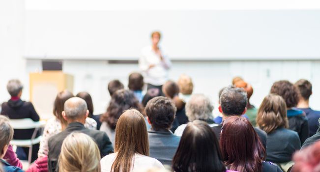 Business and entrepreneurship symposium. Female speaker giving a talk at business meeting. Audience in conference hall. Rear view of unrecognized participant in audience. Copy space on whitescreen.