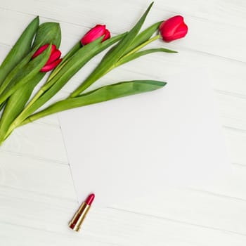 red tulips, lipstick and sheet of paper for your greetings on the background of white wooden board