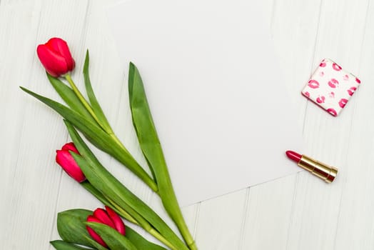 red tulips, lipstick and sheet of paper for your greetings on the background of white wooden board