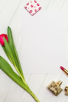 one red tulip, bottle of perfume, lipstick and sheet of paper for your greetings on the background of white wooden board