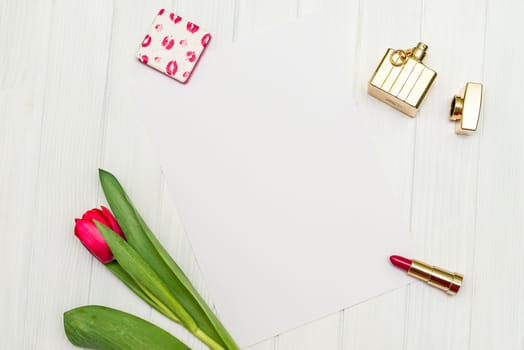 one red tulip, bottle of perfume, lipstick and sheet of paper for your greetings on the background of white wooden board