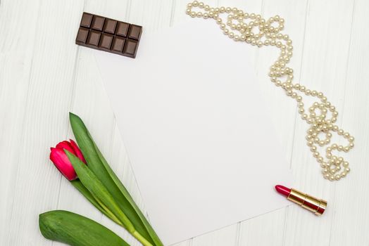 top view beautiful one red tulip, necklace, lipstick, piece of chocolate and sheet of paper for your greetings on the background of white wooden board