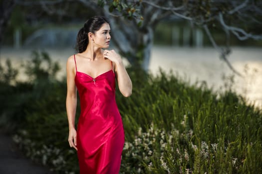 Beautiful young woman wearing a long red silk formal dress in the gardens in the afternoon.