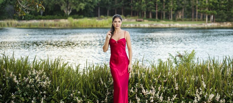 Beautiful young woman wearing a long red silk formal dress in the gardens in the afternoon. 