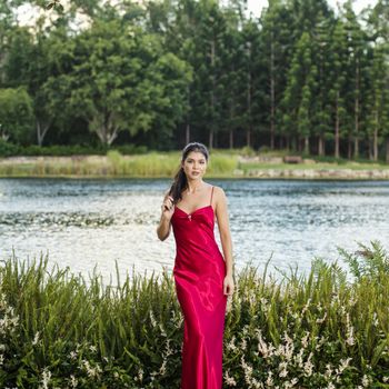 Beautiful young woman wearing a long red silk formal dress in the gardens in the afternoon