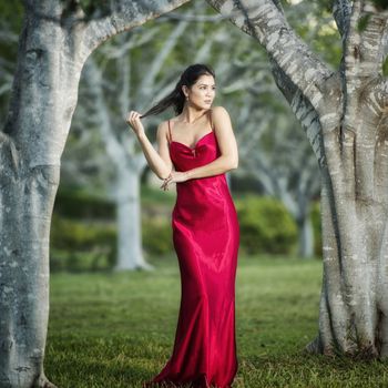 Beautiful young woman wearing a long red silk formal dress in the gardens in the afternoon. 