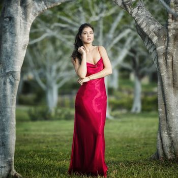 Beautiful young woman wearing a long red silk formal dress in the gardens in the afternoon.