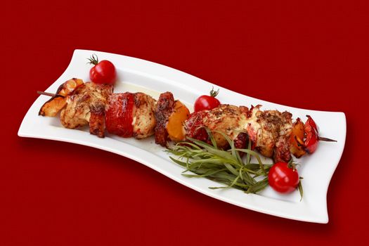 Skewers of meat and vegetables on the grill, with tomatoes and fresh herbs, on a red tablecloth