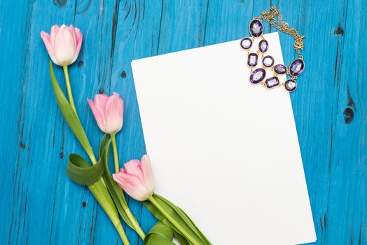 top view beautiful pink tulips, necklace and sheet of paper for your greetings on the background of blue wooden board