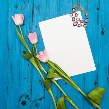 top view beautiful pink tulips, necklace and sheet of paper for your greetings on the background of blue wooden board