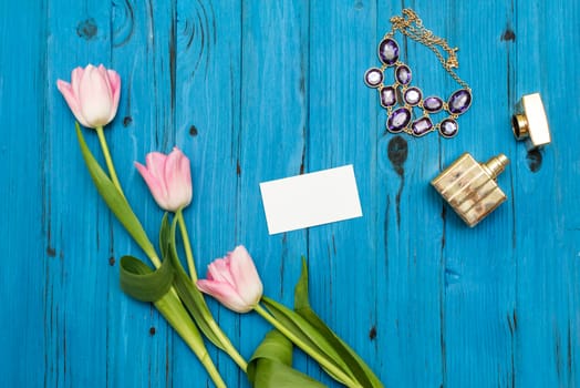 top view beautiful pink tulips, necklace, bottle of perfume and sheet of paper for your greetings on the background of blue wooden board