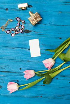 top view beautiful pink tulips, necklace, bottle of perfume and sheet of paper for your greetings on the background of blue wooden board