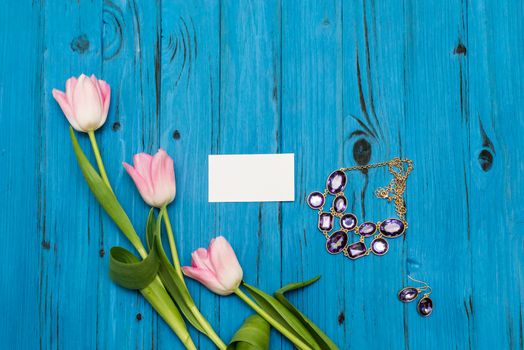 top view beautiful pink tulips, necklace with earring and card of paper for your greetings on the background of blue wooden board