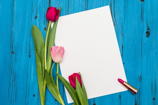 top view of red and pink tulips, lipstick and sheet of paper for your greetings on the background of blue wooden board
