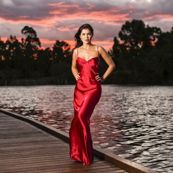 Beautiful young woman wearing a long red silk formal dress in the gardens in the afternoon. 