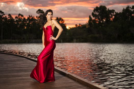 Beautiful young woman wearing a long red silk formal dress in the gardens in the afternoon.