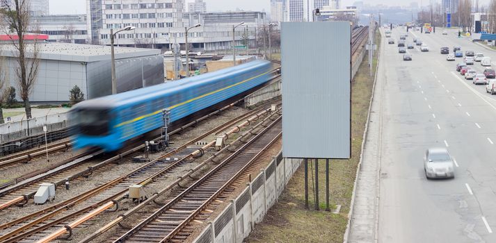 Train of a metro system moving on the overground part of the subway line and city street with moving cars beside 
