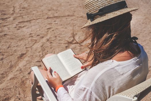 Young adult woman with a hat on the beach reading a book.