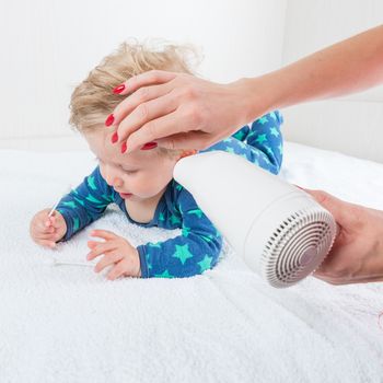 Mom dries baby boy hair with a hair dries, while the infant toys lying with cotton swab.