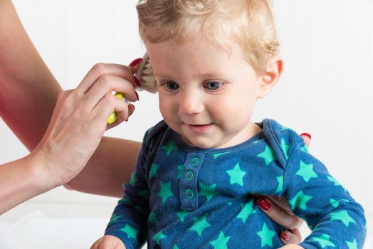 Mom combing the baby with blonde hair, while the toddler is sitting.