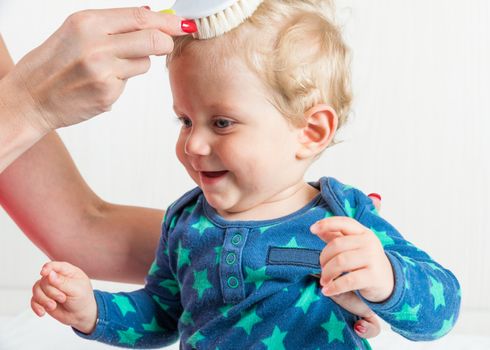 Mom combing the baby with blonde hair, while the toddler is sitting.