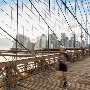 Blured runner running across Brooklyn bridge. New York City Manhattan downtown skyline in sunset with skyscrapers illuminated over East River panorama as seen from Brooklyn bridge.