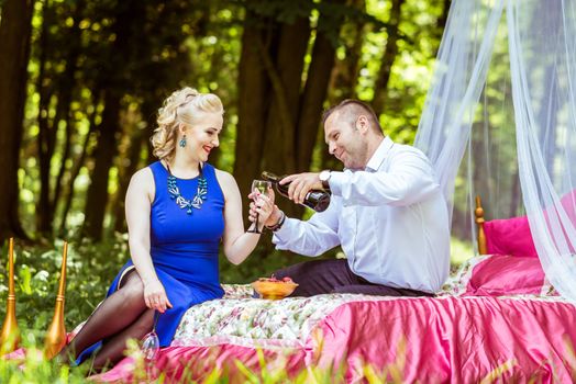 Man and woman sitting on the bed in the lawn and man pours wine from a bottle into a glass in Lviv, Ukraine.