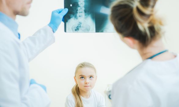 Little Girl Awaiting Medical Diagnosis in Front of Two Hospital Physicians with X Ray Image.