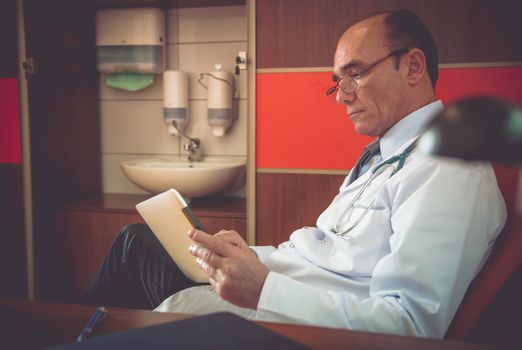Senior Caucasian Medical Doctor in the Office Browsing Data on His Private Tablet Computer. 