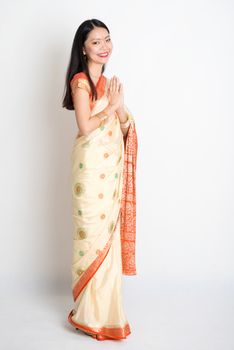 Full length mixed race Indian Chinese girl with sari dress in greeting gesture, standing on plain background.