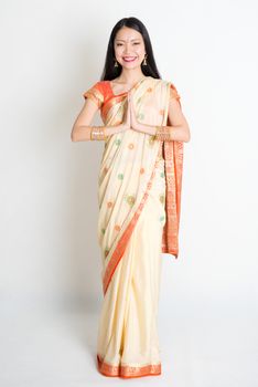 Full length mixed race Indian Chinese female with sari dress in greeting gesture, standing on plain background.