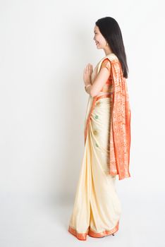 Full length side view mixed race Indian Chinese female with sari dress in greeting gesture, standing on plain background.
