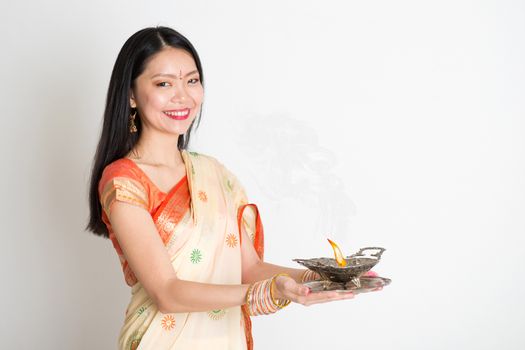 Portrait of young mixed race Indian Chinese female in traditional sari dress, holding diya oil lamp, on plain background.