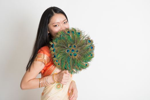 Portrait of young mixed race Indian Chinese female in traditional sari dress, holding peacock feathers fan covering face, on plain background.