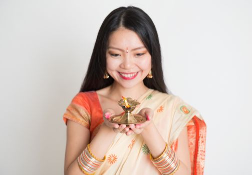 Portrait of young mixed race Indian Chinese girl in traditional sari dress, holding diwali oil lamp light, on plain background.