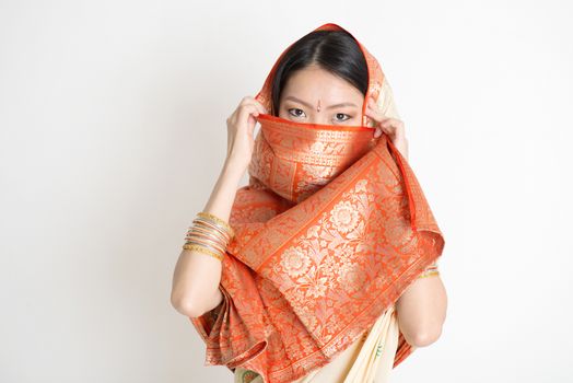 Portrait of secrecy mixed race Indian Chinese woman in traditional sari dress, covering face and looking at camera, on plain background.