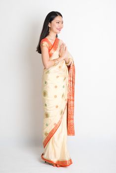 Full length mixed race Indian Chinese woman with sari dress in greeting gesture, standing on plain background.