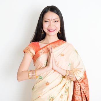 Portrait of mixed race Indian Chinese girl with traditional sari dress in greeting gesture, standing on plain background.