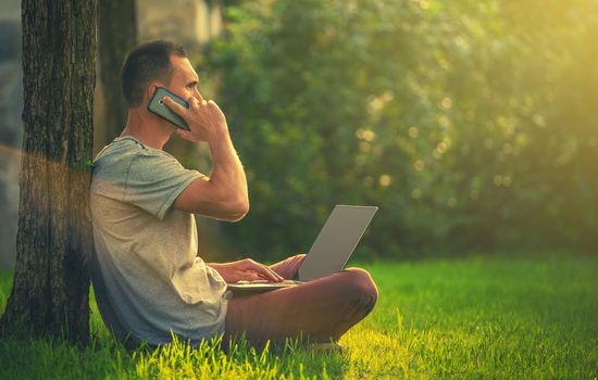 Outdoor Office Worker. Men Making Business Calls While Seating on the Grass in a Park with Laptop Computer on His Legs. Out of Office Work.