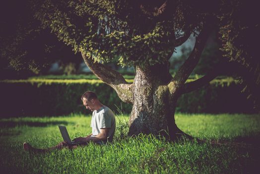 Internet Break Under the Tree. Men Enjoying Wireless Internet Connectivity. WiFi Connection in the Garden. Caucasian Men Working Remotely on His Laptop Computer.