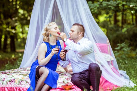 Man and woman sitting on the bed in the lawn with glasses of wine and woman eating strawberries from a man's hand in Lviv, Ukraine.
