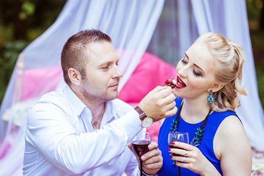 Man and woman sitting on the bed in the lawn with glasses of wine and woman eating strawberries from a man's hand in Lviv, Ukraine.